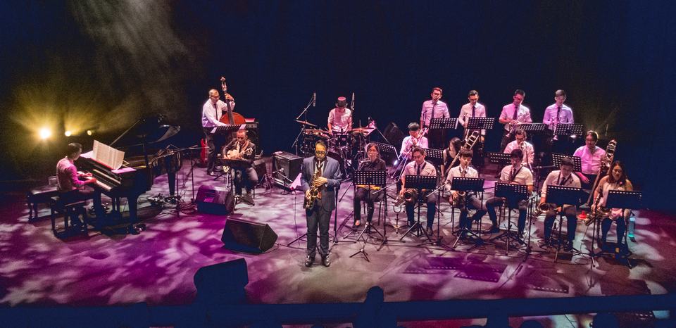 The Julian Chan Jazz Orchestra on a theatre stage: piano, bass, drums and guitar on the left, the saxophone, trombone and trumpet sections seated behind music stands, and a saxophonist standing at the front.